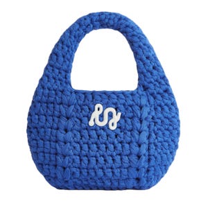 A blue, handcrafted crocheted handbag with a textured design and a short handle. It features a white, wavy logo or symbol on its front.