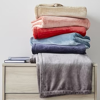 A stack of folded plush blankets in various colors is displayed on a wooden dresser.
