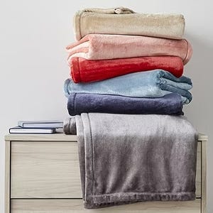 A stack of folded plush blankets in various colors is displayed on a wooden dresser.