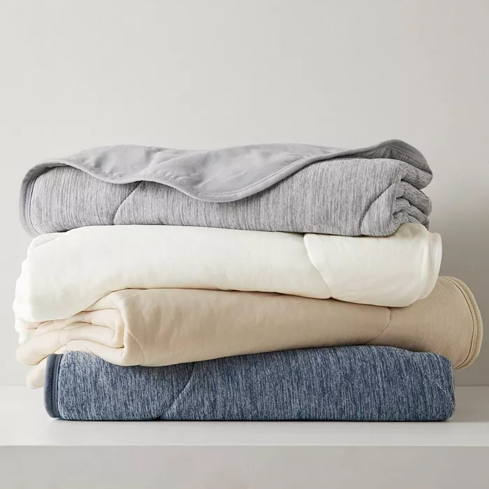 Four folded blankets in varying shades of gray, cream, beige, and blue are stacked neatly on a white surface.