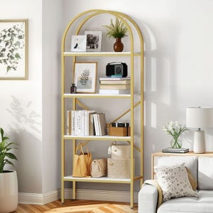 A tall, gold-piped bookshelf with five shelves holds books, a radio, framed photos, a vase with greenery, decorative boxes, and handbags. A nearby small table has a lamp and flowers, and artwork is displayed on the wall.