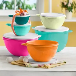 A set of colorful plastic mixing bowls with lids in various sizes is displayed, with a small teal bowl adorned with an orange ribbon and flowers on top. Three eggs and a folded kitchen cloth are placed next to the orange bowl.