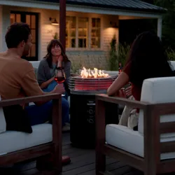 A group of people is sitting around a Ninja outdoor woodfire grill, enjoying drinks on a patio with string lights and a house in the background.
