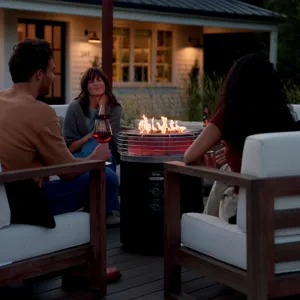 A group of people is sitting around a Ninja outdoor woodfire grill, enjoying drinks on a patio with string lights and a house in the background.