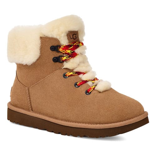 The UGG Alpine Mini Boot features a tan suede exterior, white plush shearling lining, and colorful red and yellow laces with black eyelets. It has a sturdy brown sole and a fold-over cuff for added warmth and style.