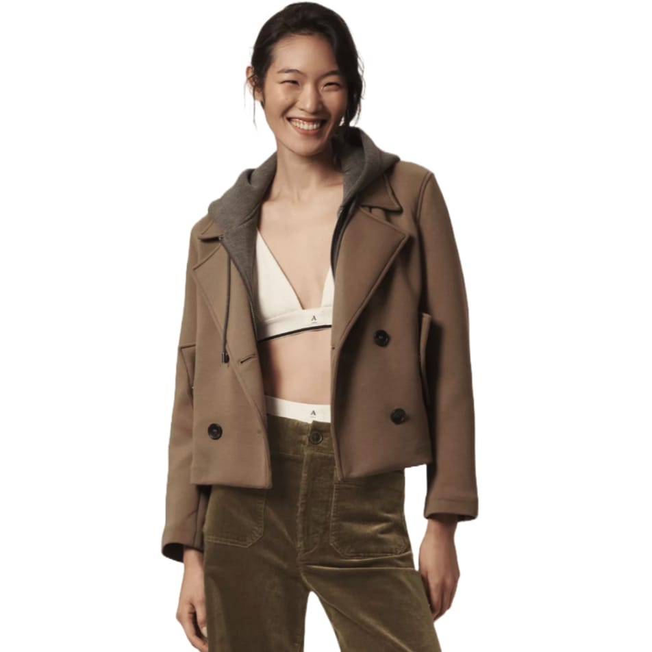 Tan hooded peacoat with a double-breasted design, featuring black buttons and a gray hoodie insert. Worn over a white bralette and paired with high-waisted olive pants.